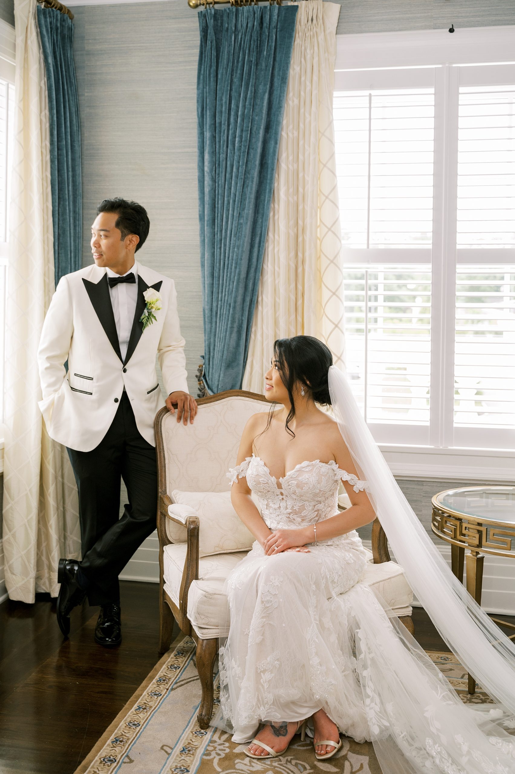 bride sits on white chair while groom stands behind her looking out window
