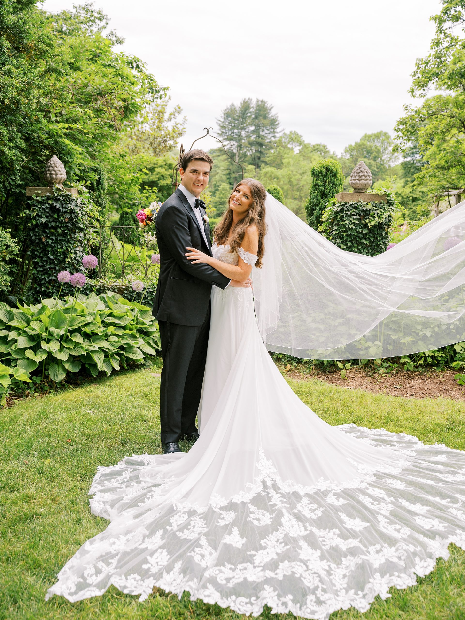 groom in black tux hugs bride with her veil floating in garden at Willowwood Arboretum