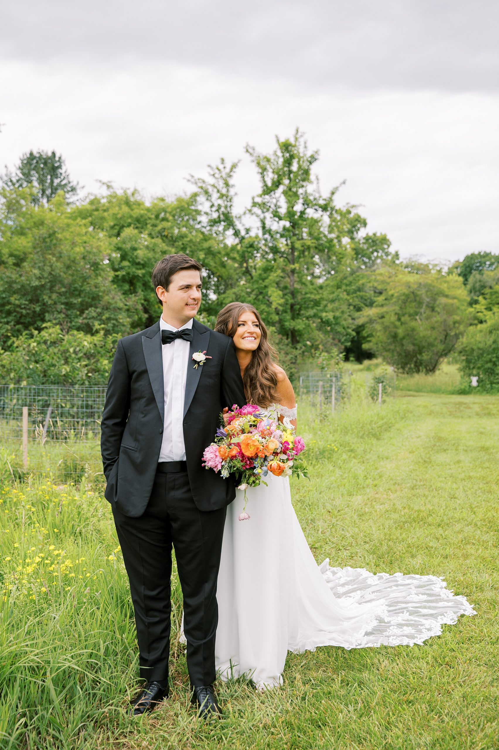 bride in off-the-shoulder gown stands slightly behind groom in black tux in field with bright flowers 