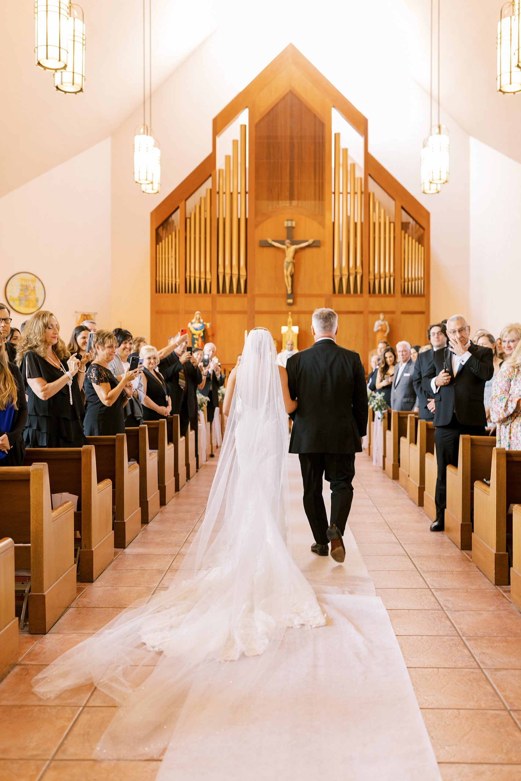 bride walks down aisle with dad with veil trailing behind her during traditional Catholic Church wedding at St. Augustine Catholic Church