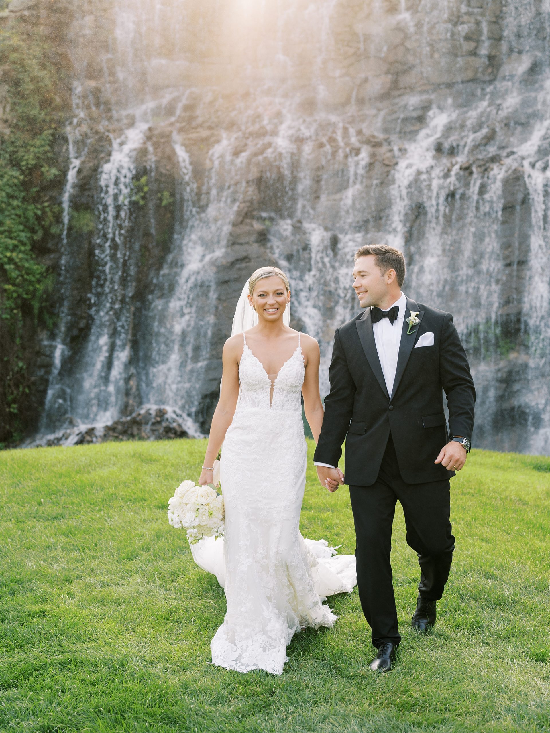 bride and groom hold hands walking away from waterfall