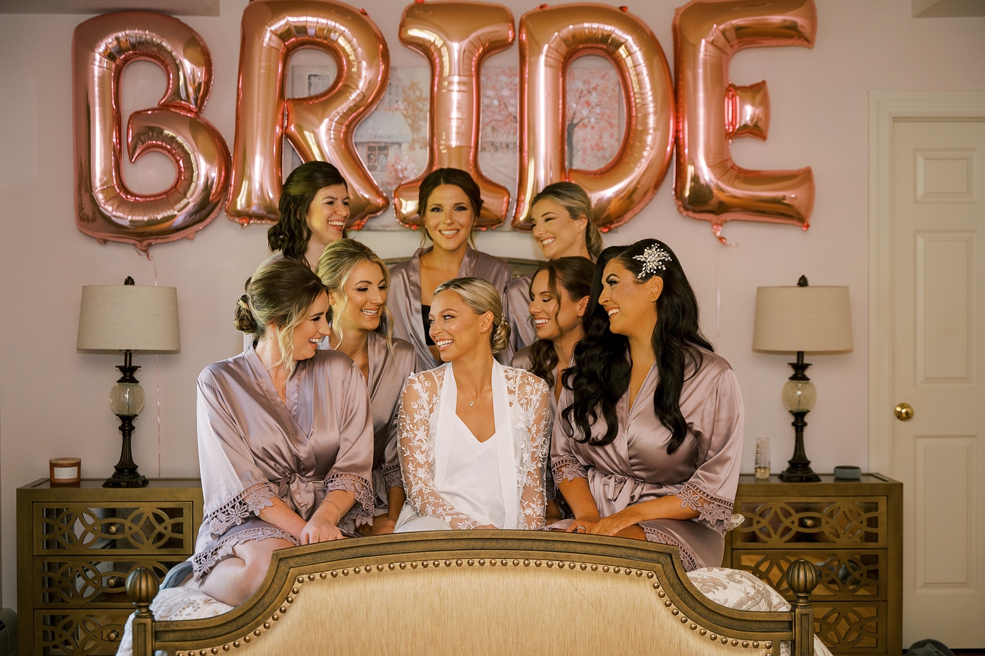 bride sits with bridesmaids under "BRIDE" balloons on bed
