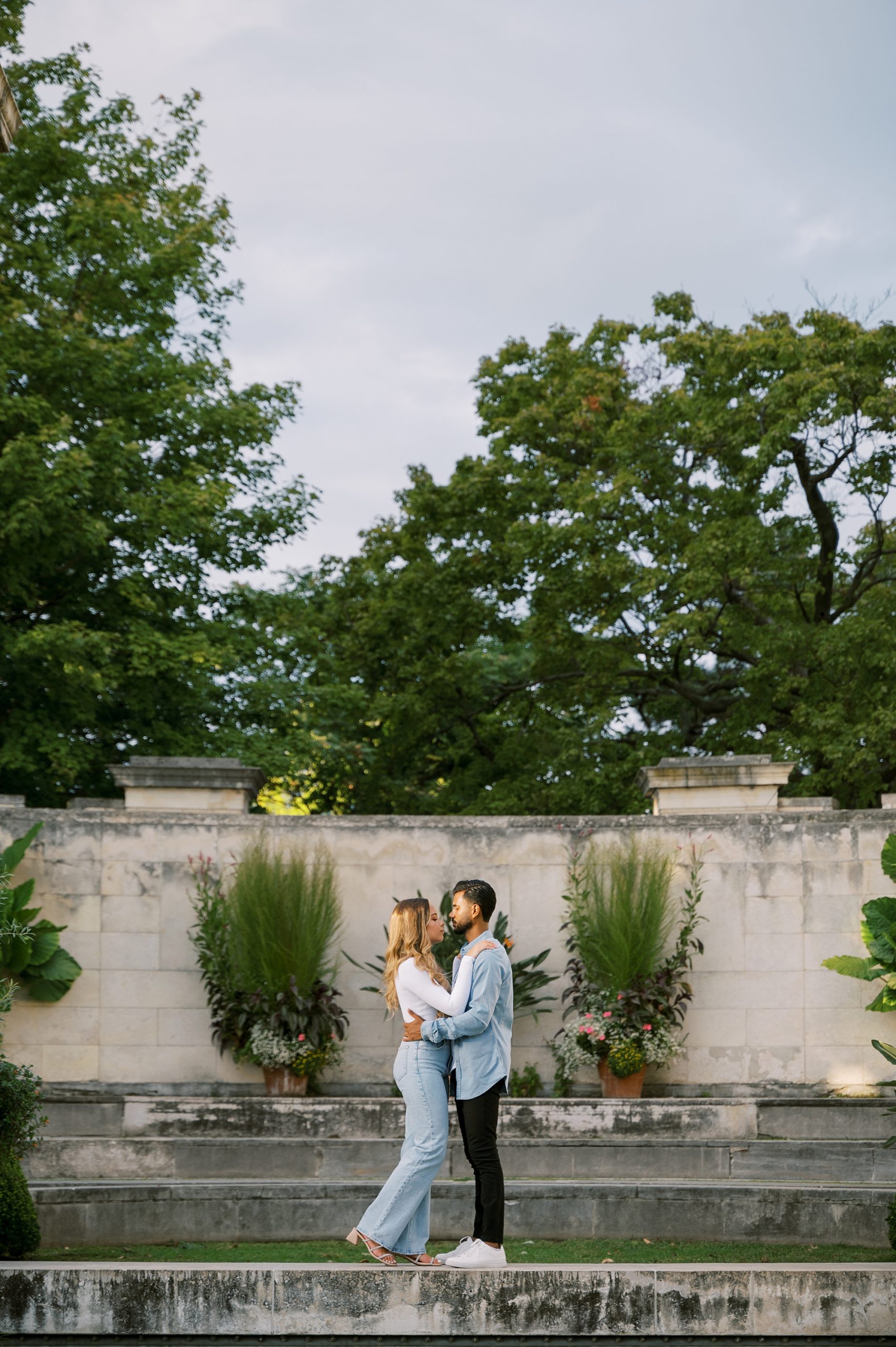 engaged couple in casual outfits kiss on stone steps inside Untemyer Gardens Conservancy