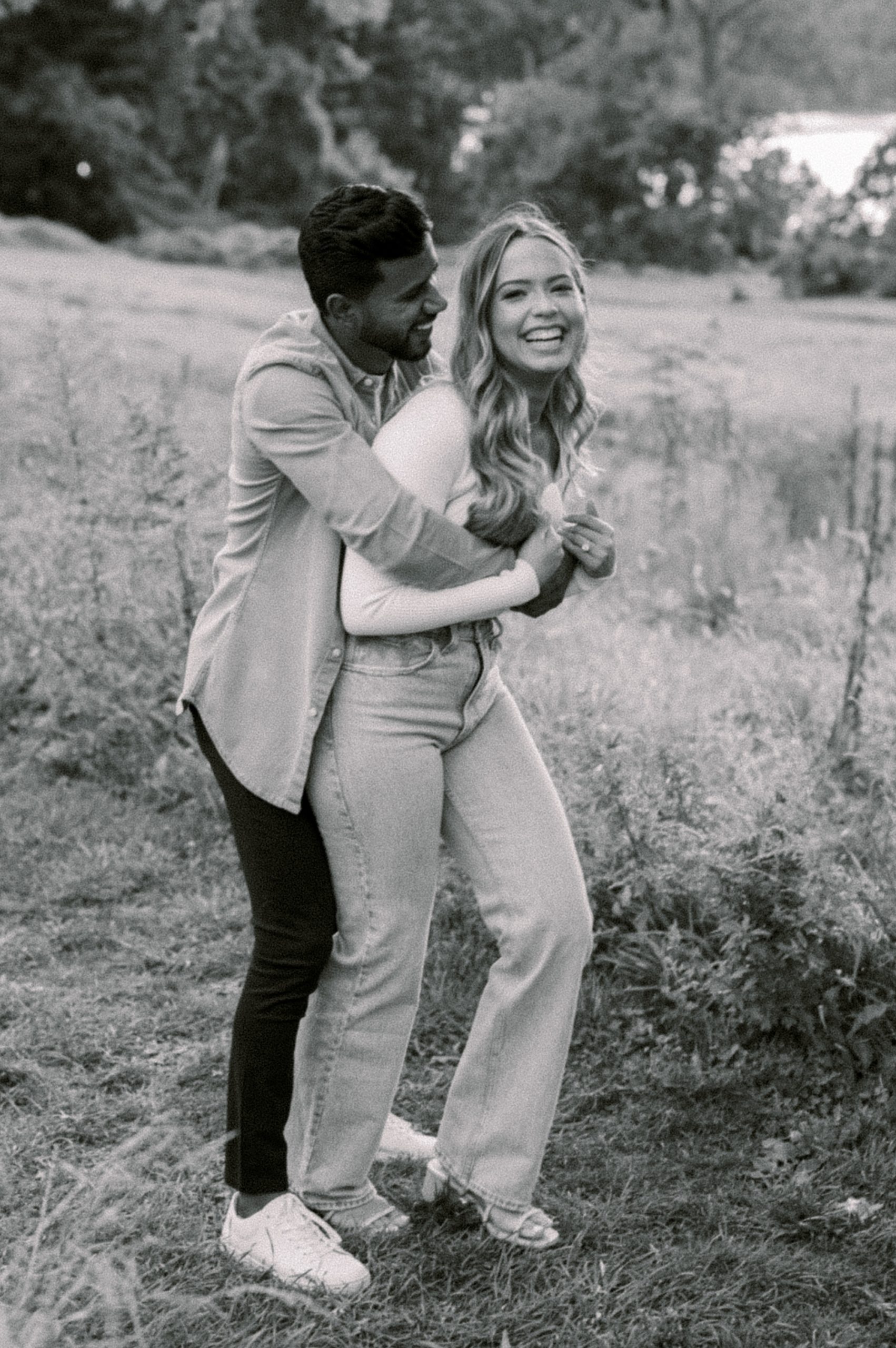 man hugs fiancee from behind making her laugh in field