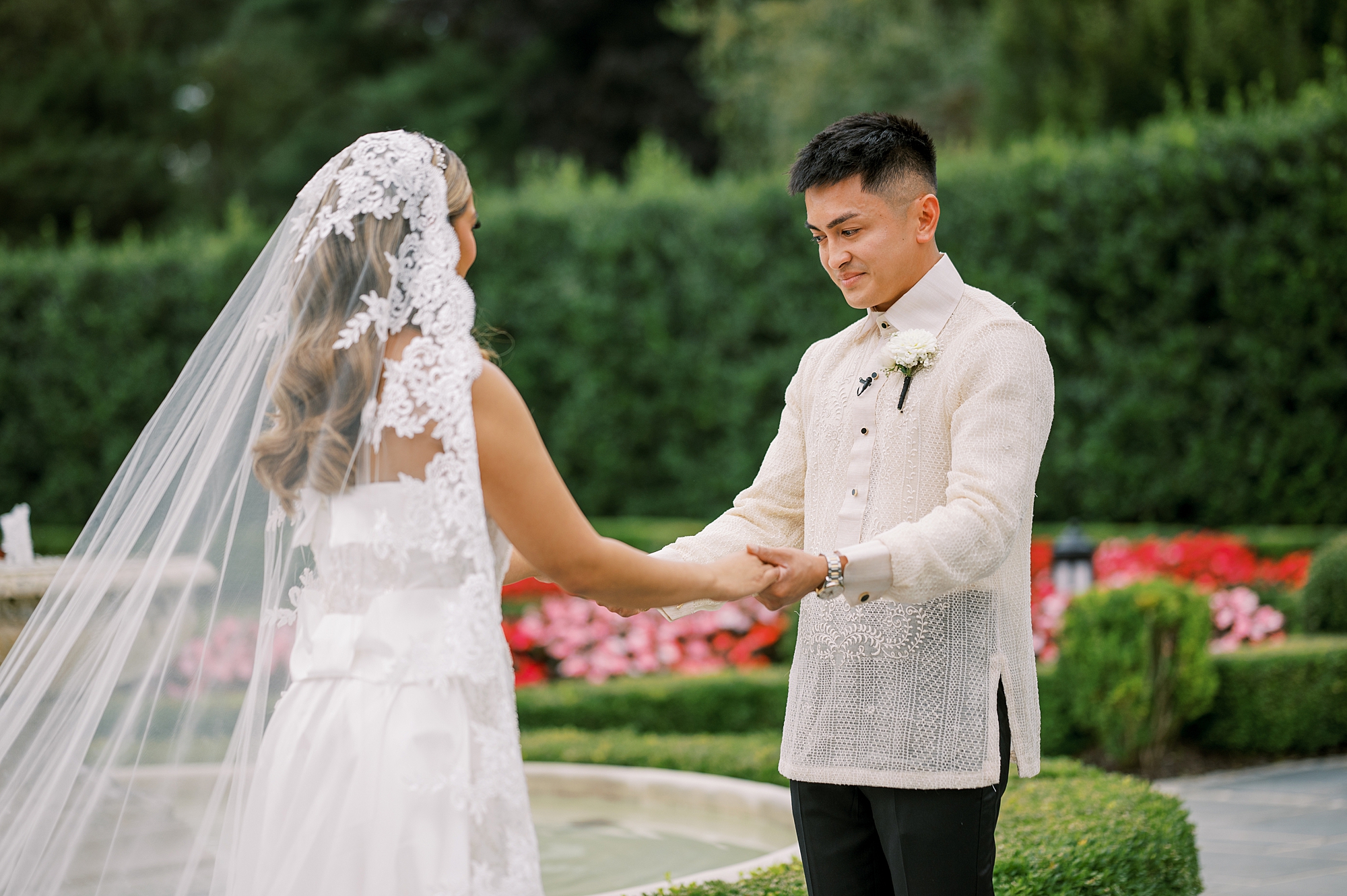 groom holds bride's hand looking at her wedding dress during first look in gardens of Park Chateau Estate