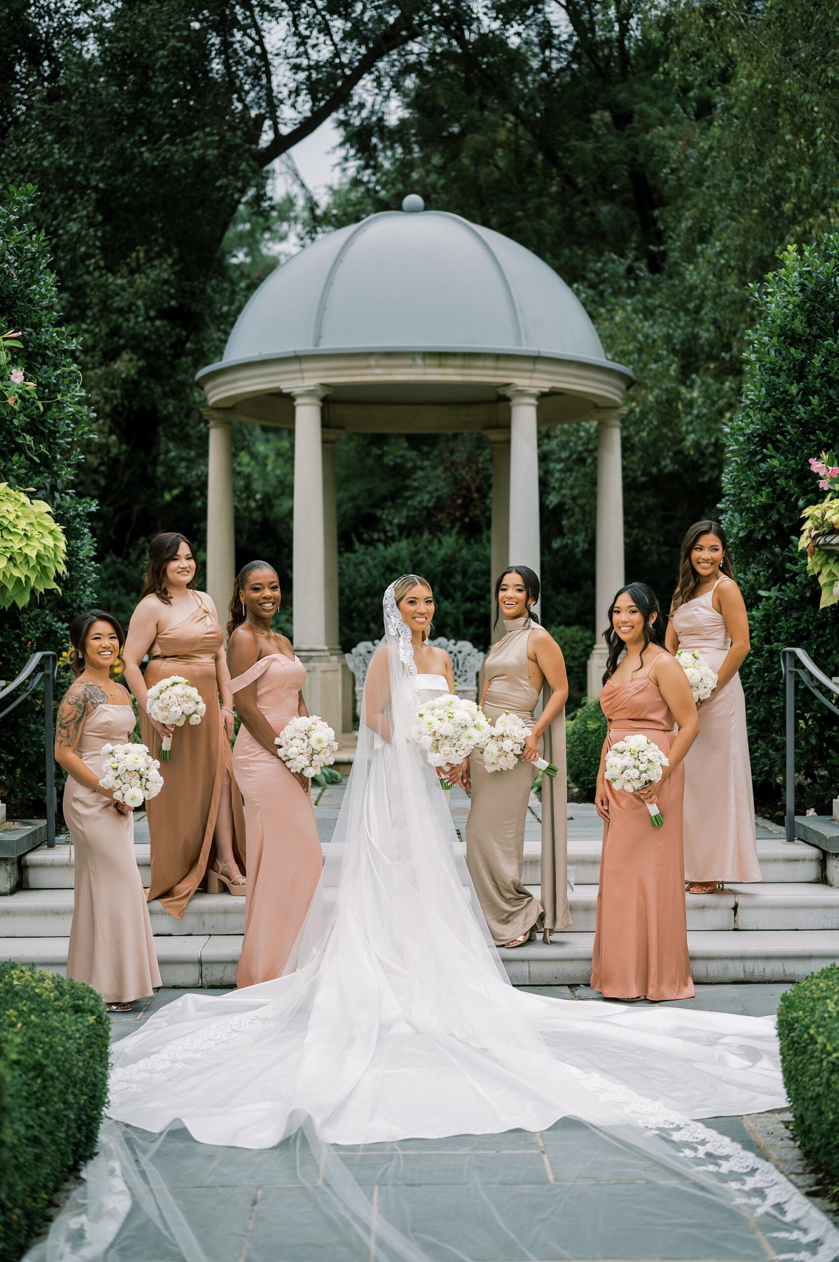 bride stands in front of gazebo at Park Chateau with bridesmaids in pink gowns