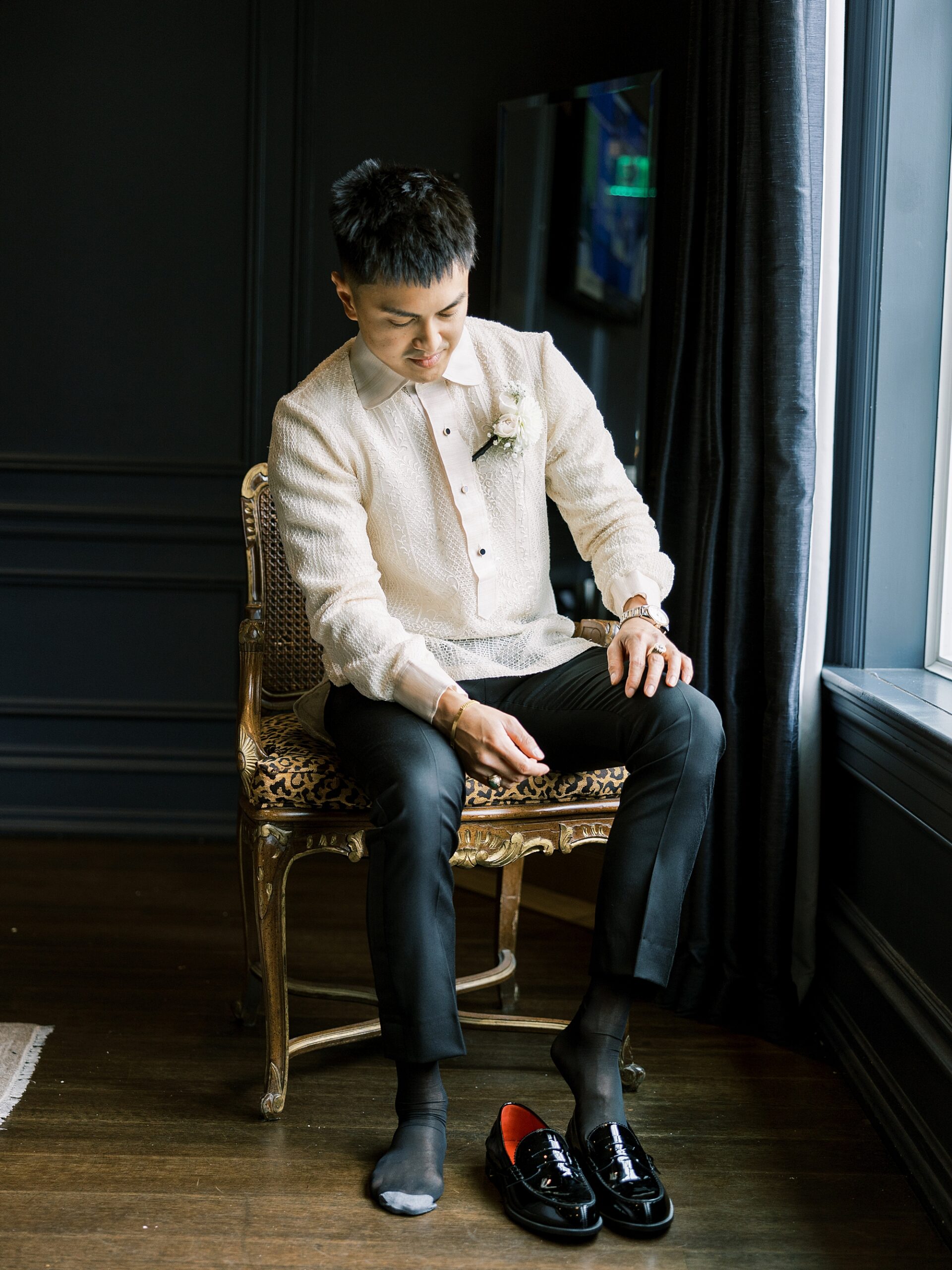 groom sits by window putting on black shoes 