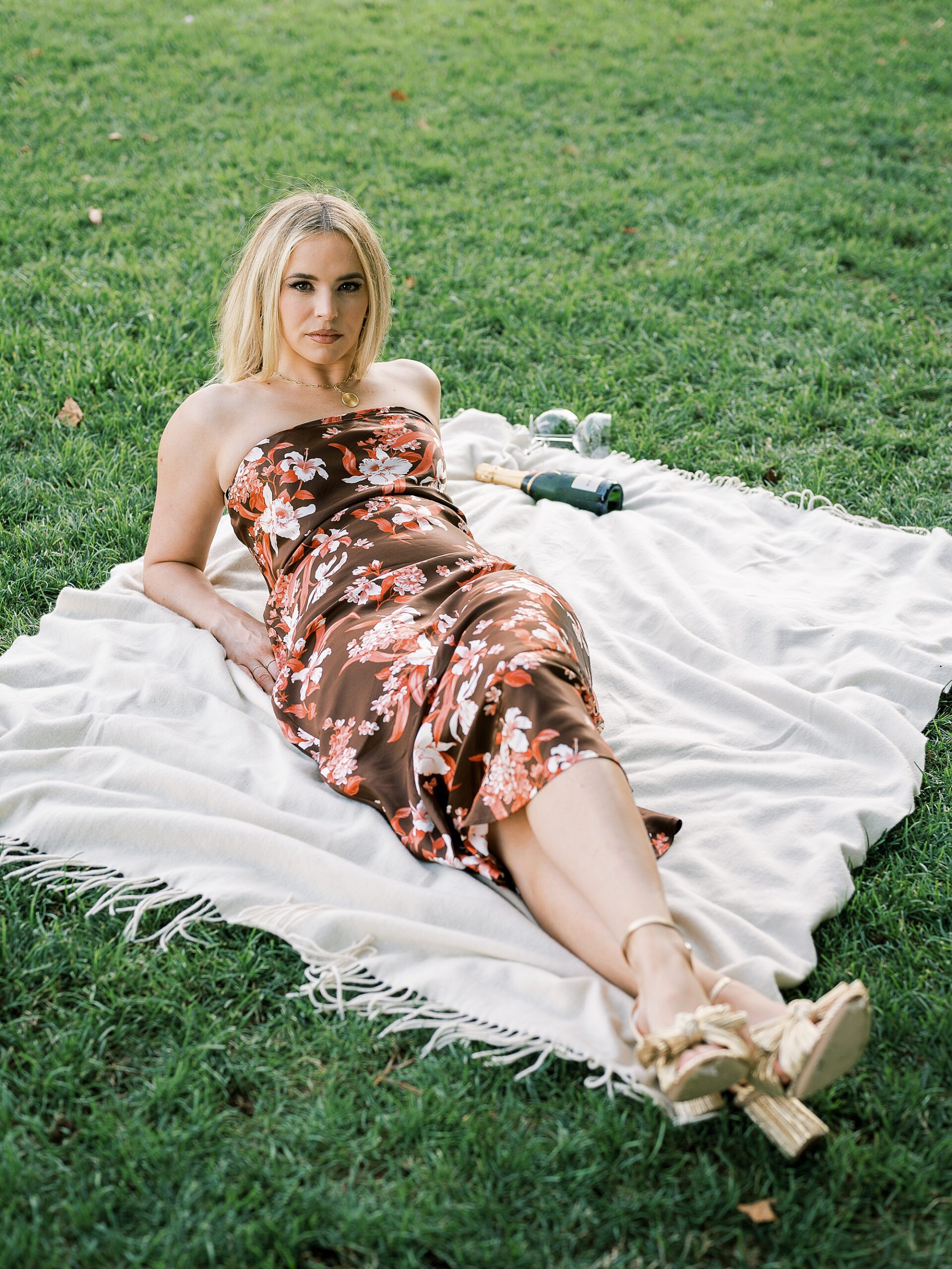 blonde woman in brown floral dress lays back on white picnic blanket 