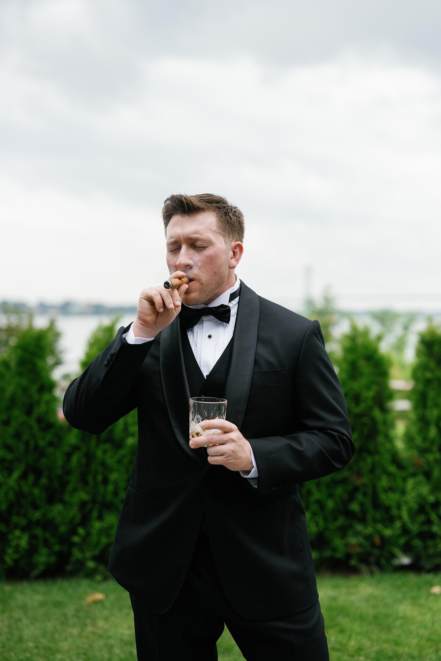 groom holds glass of whiskey and smokes cigar in front of trees