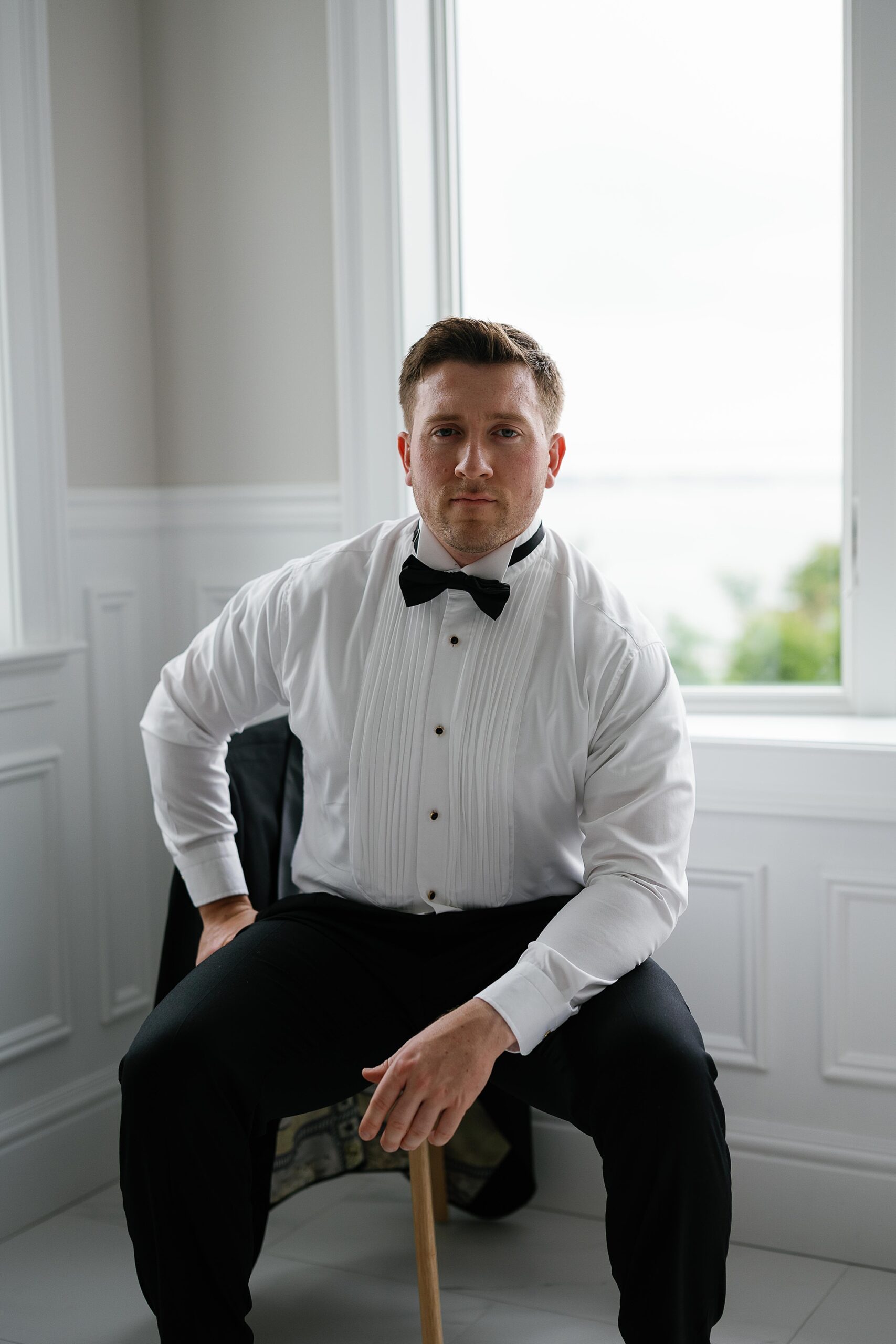 groom sits in corner of room in white shirt and black tie