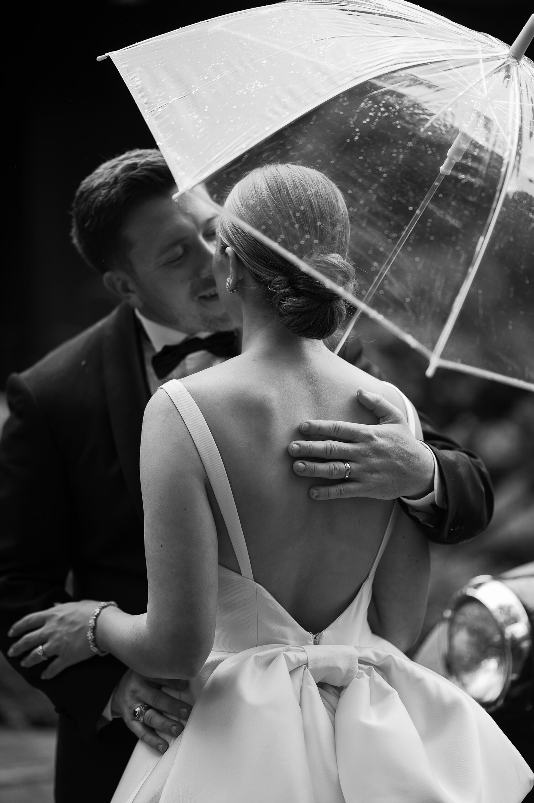 groom leans to kiss bride under clear umbrella on rainy wedding day