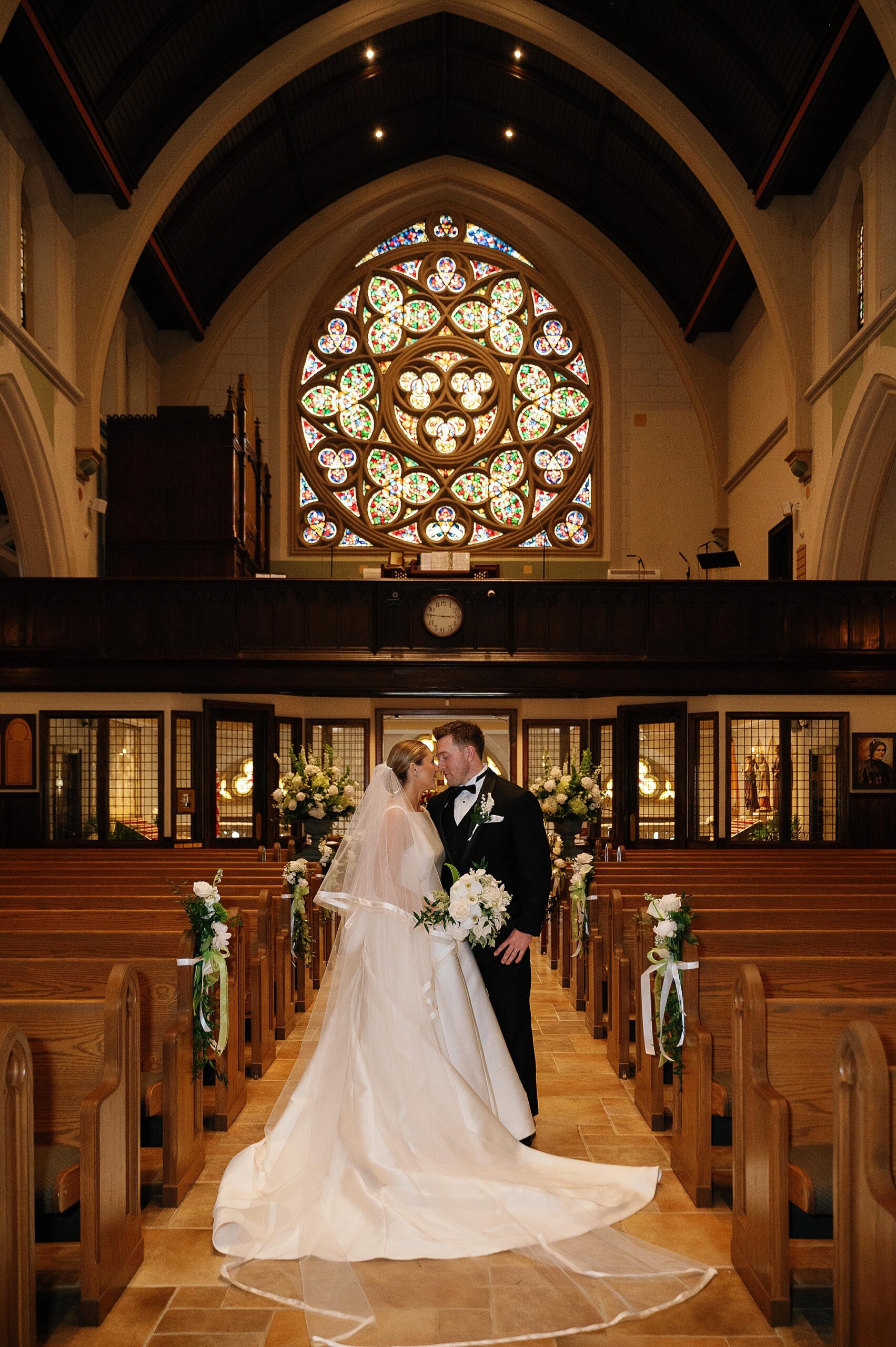 bride and groom kiss in aisle of wooden pews in front of stained glass window at Immaculate Conception Church