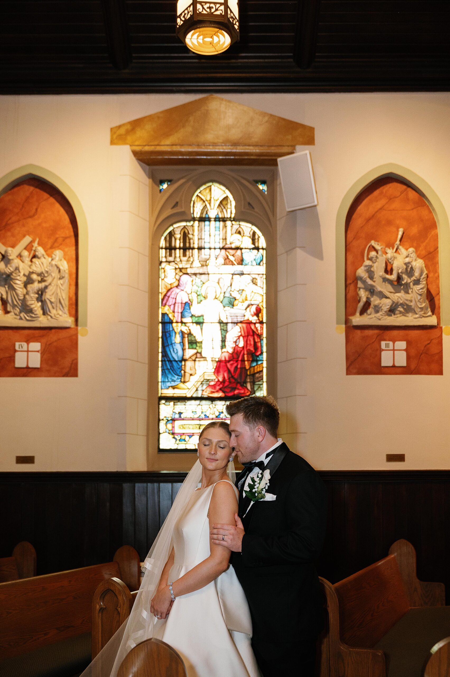 groom hugs bride from behind standing between wooden pews at Immaculate Conception Church