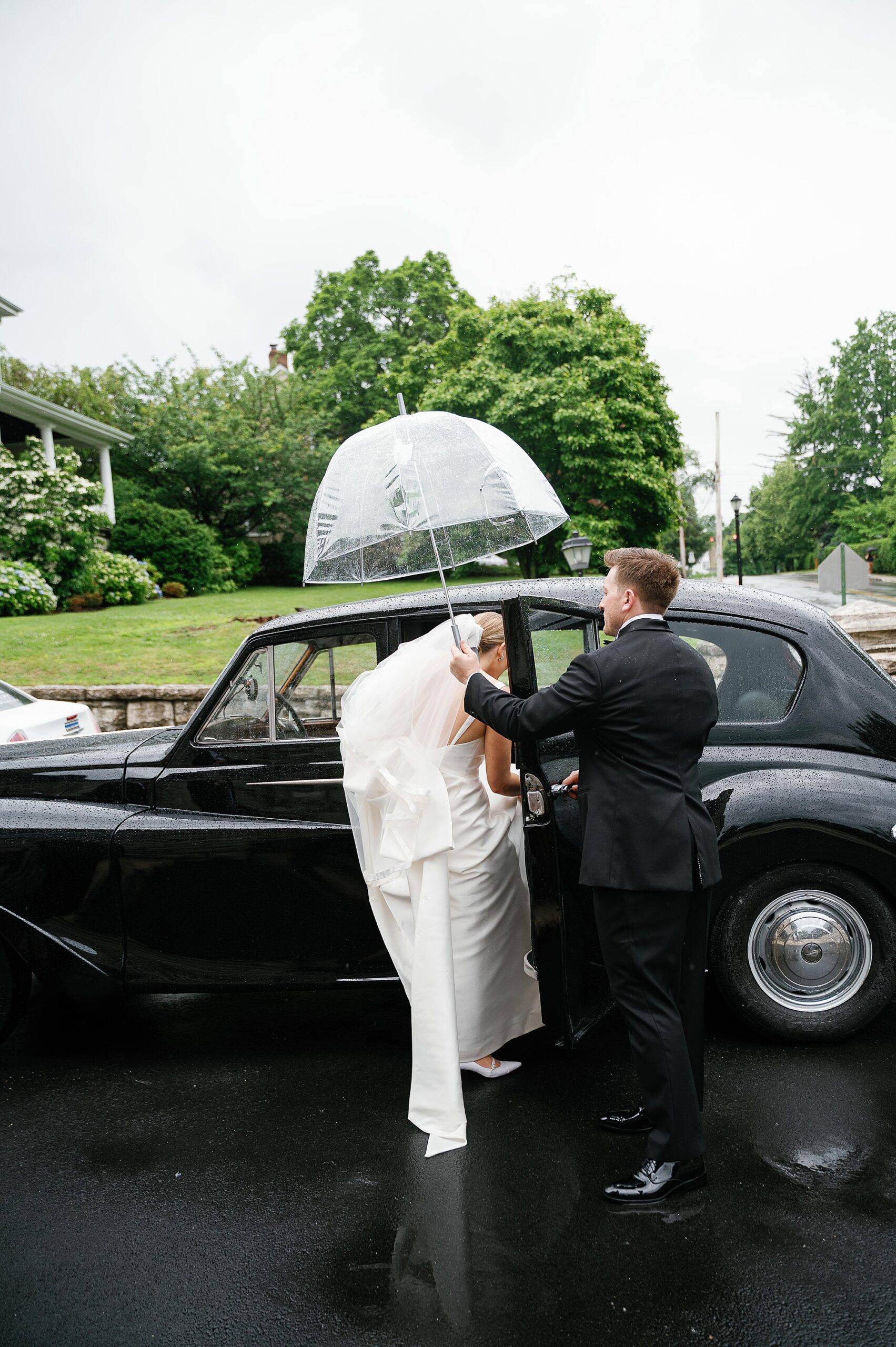 groom holds umbrella over bride as she steps into black classic car